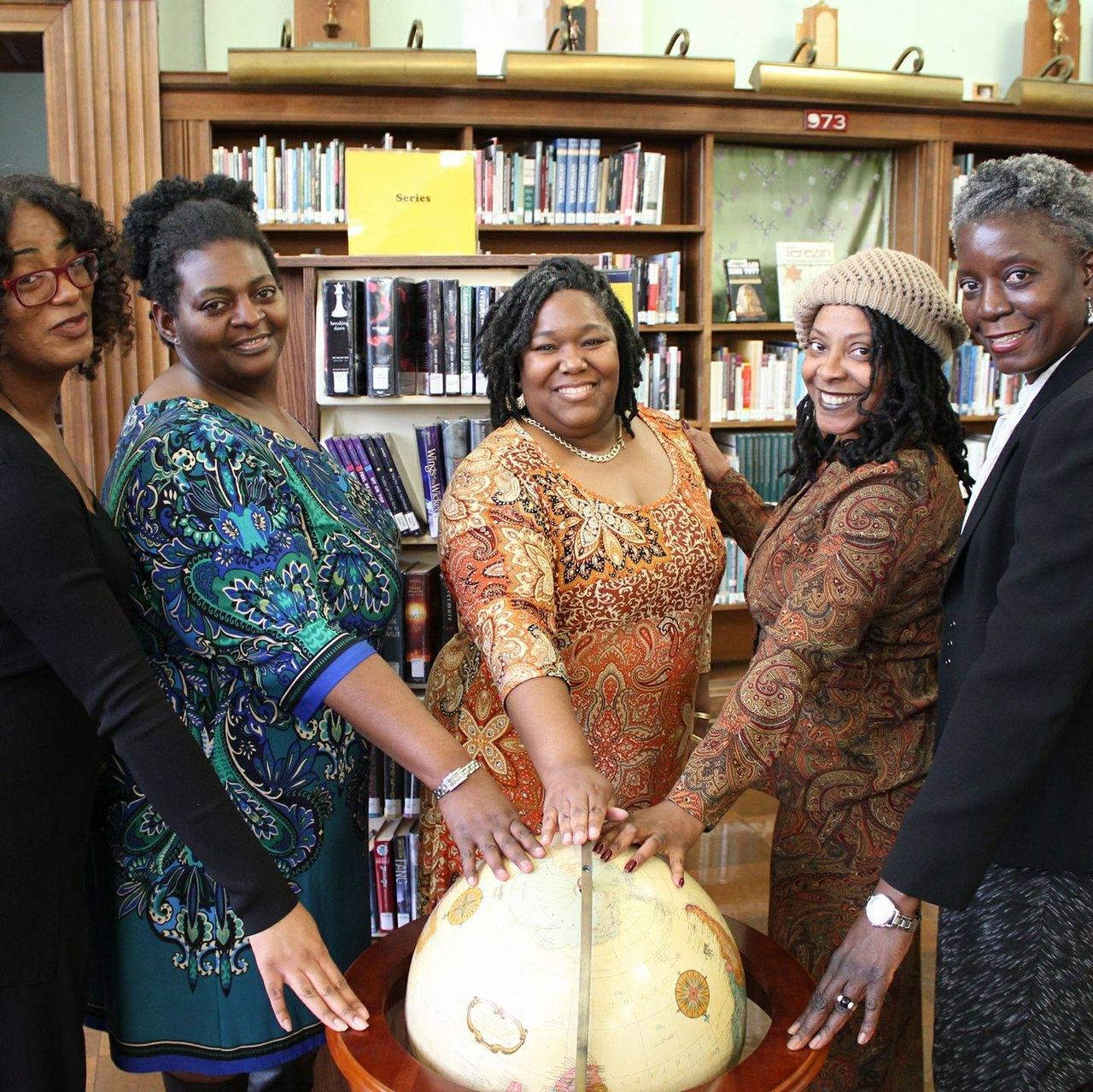 Five women gather around a globe in a library, symbolizing unity and education.